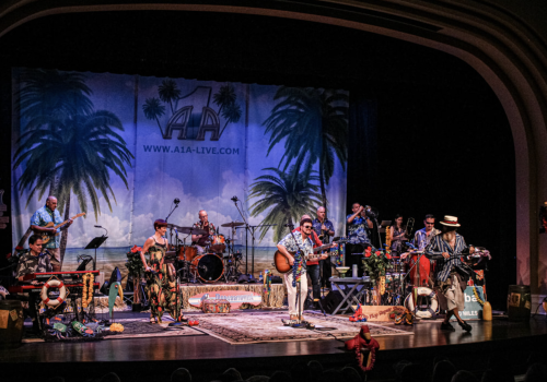 A band performs on a decorated tropical-themed stage with instruments and various props under an 