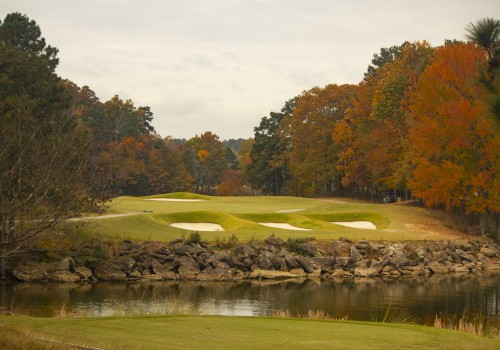 A scenic golf course with manicured greens, sand bunkers, a rock wall, and a water hazard, surrounded by autumn foliage and a cloudy sky.