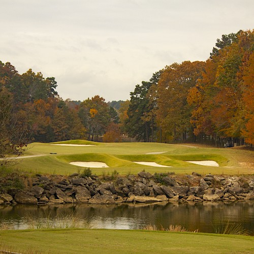 A scenic golf course with manicured greens, sand bunkers, a rock wall, and a water hazard, surrounded by autumn foliage and a cloudy sky.