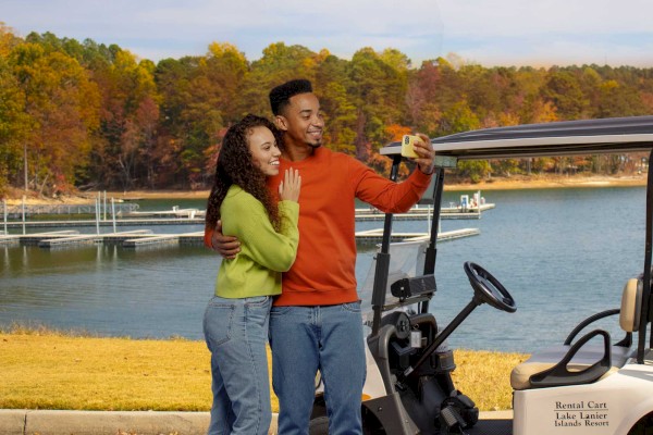 A couple is taking a selfie by a golf cart near a lake, with autumn trees in the background.