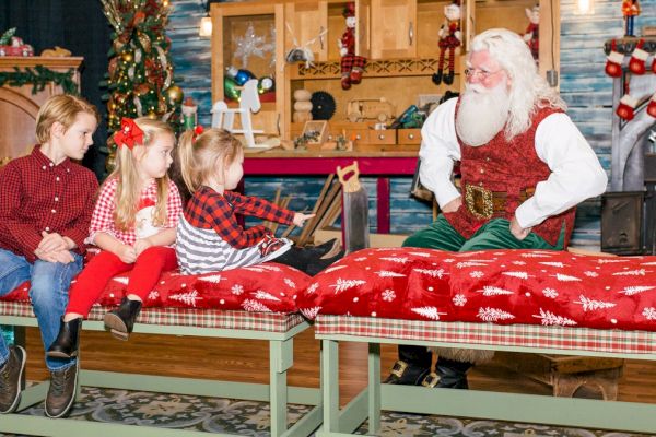 Four children are sitting on benches, talking to a man dressed as Santa Claus in a festive, holiday-themed setting.