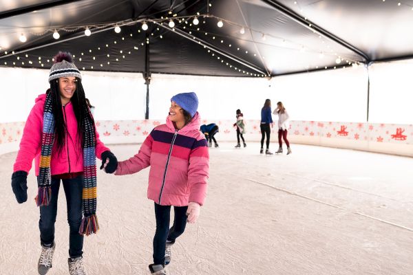 People ice skating in an indoor rink with string lights overhead, wearing winter clothing and smiling, enjoying the activity.