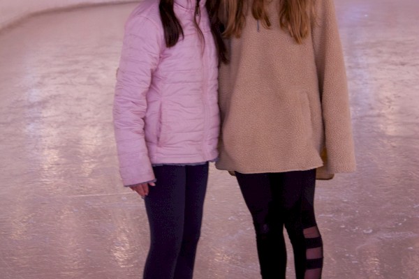 Two girls pose together on an ice skating rink, each wearing winter clothes and ice skates, smiling for the camera.