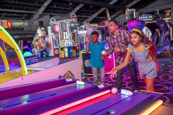 Three people are enjoying games in an arcade. One girl is actively rolling a ball in a skee-ball game while two boys watch and cheer her on.