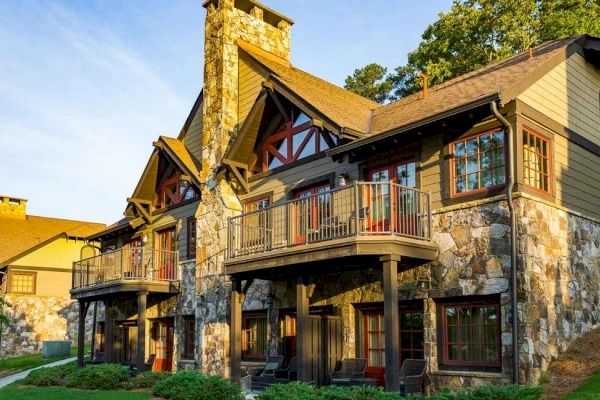 A rustic, multi-story building with stone and wood accents, featuring balconies and a chimney, surrounded by greenery under a clear sky.