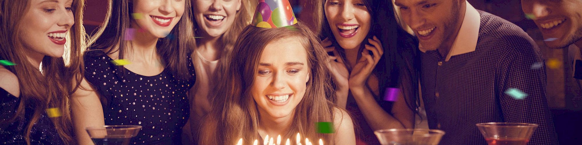 A group of people wearing party hats gather around a woman about to blow out candles on a birthday cake. Confetti falls around them.