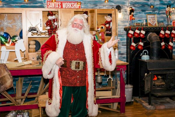 A person dressed as Santa Claus stands in a festive workshop setting, complete with Christmas decorations and a 