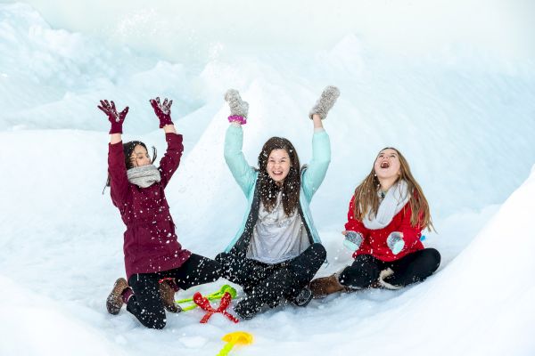 Three people are sitting in the snow, throwing snow into the air, and smiling joyfully. Brightly colored toys are scattered nearby.