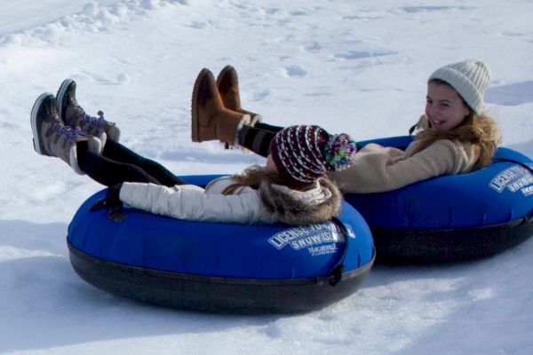 Two people wearing hats and winter clothing are snow tubing on blue inflatable tubes on a snowy slope, smiling and enjoying the activity.