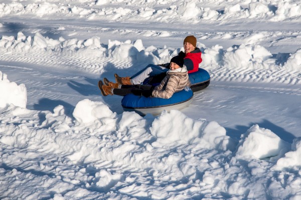 Two people are tubing down a snowy slope, wearing winter clothing and enjoying the outdoor activity.