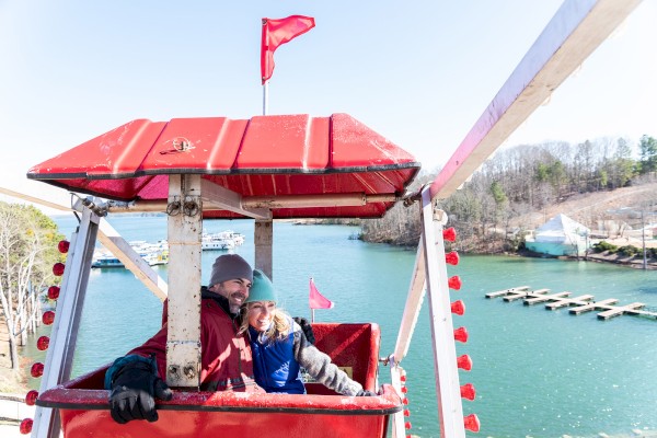 Two people are enjoying a ride in a red Ferris wheel cabin by a serene lake, with a picturesque backdrop of trees and docks ending the sentence.