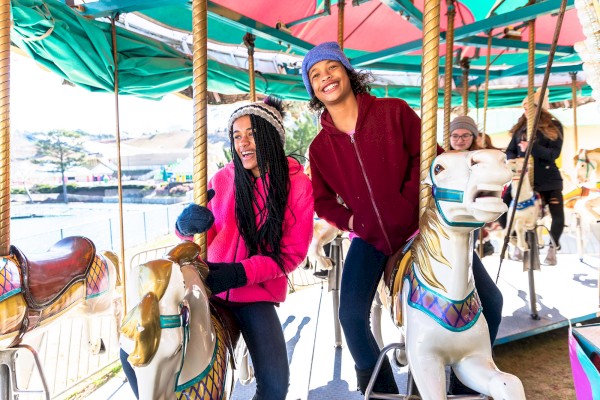 Two people are riding a carousel, smiling and enjoying themselves, with others in the background also on the ride.