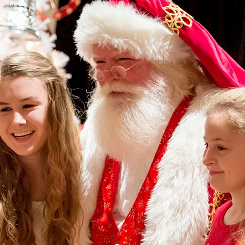 Two young girls are posing with a man dressed as Santa Claus in front of a festive Christmas tree, all smiling happily.