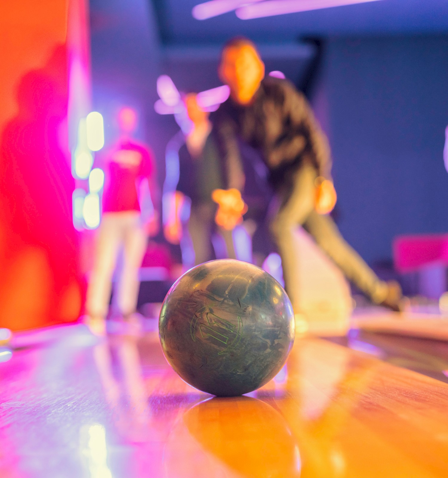 People are bowling in a neon-lit alley with an out-of-focus background and a bowling ball in the foreground.