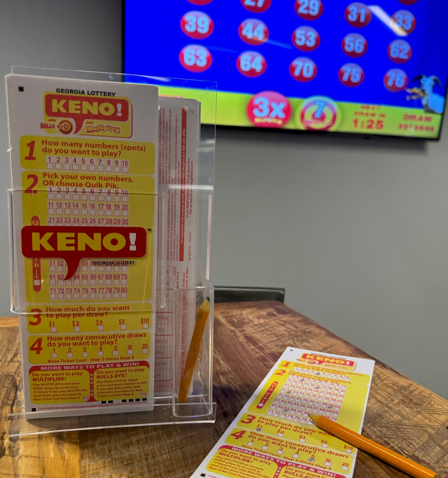 The image shows Keno lottery tickets and a pencil on a table, with a Keno numbers display screen in the background.