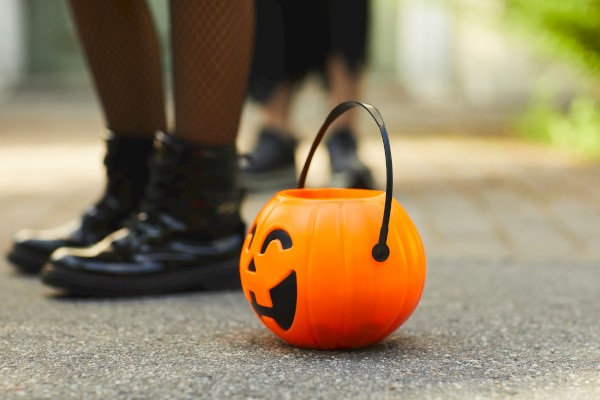 A small orange pumpkin-shaped bucket with a smiling face on the ground, with people in Halloween costumes standing in the background.