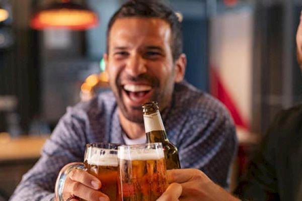 A group of friends share a happy moment, clinking their beer bottles and glasses together in a lively setting while smiling and laughing.