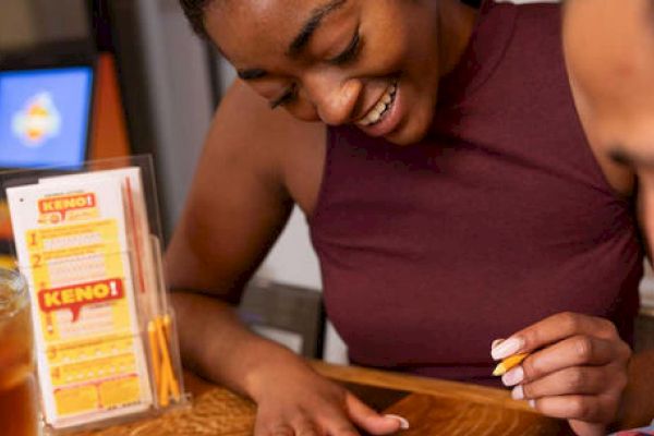 Two people sitting at a table, with one holding a pencil, possibly playing Keno. A drink and a Keno pamphlet with pencils are on the table.