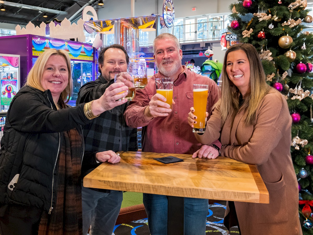 Four people are raising glasses in a toast while standing around a wooden table, with a Christmas tree and arcade games in the background.