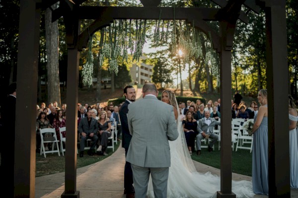 A wedding ceremony taking place outdoors under a wooden arch with greenery, a couple facing the officiant, and guests seated watching.