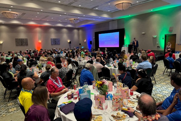 People are gathered in a large conference room with decorated tables, enjoying a meal while attending a presentation on a stage.