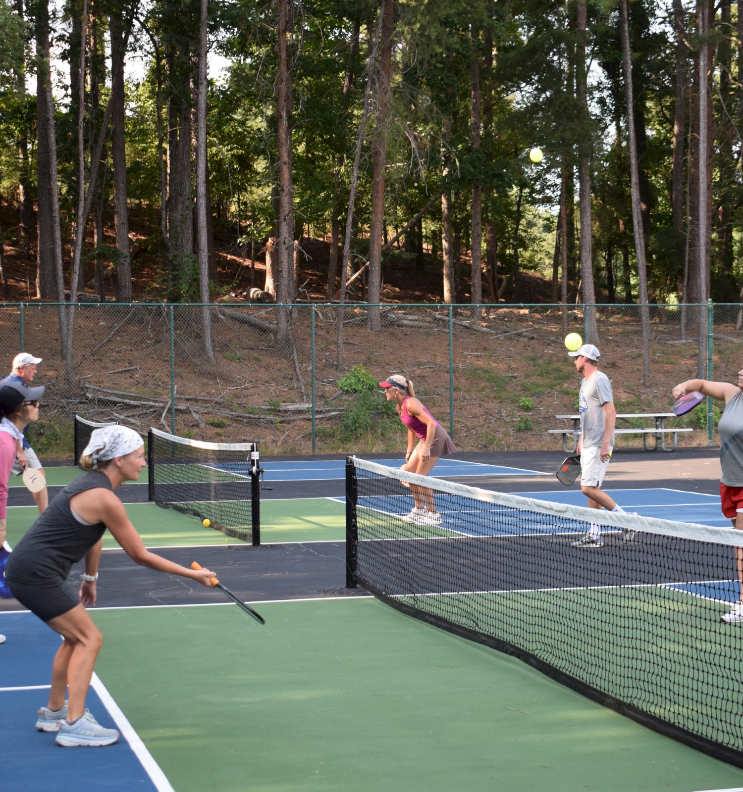 People are playing pickleball on an outdoor court with trees in the background.