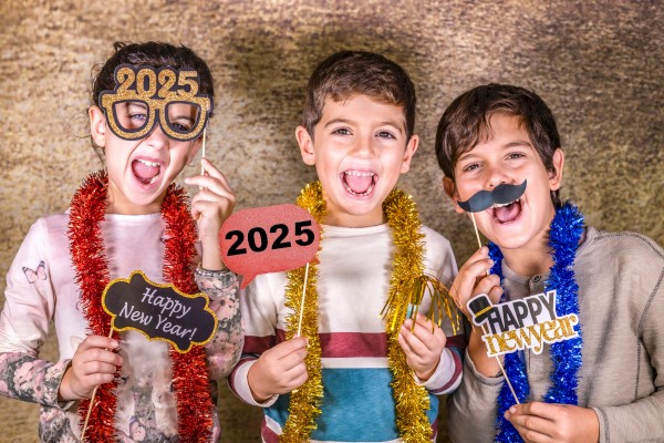 Three children celebrating New Year 2025 with props like glasses, mustaches, and signs. They are adorned with red and gold tinsel.