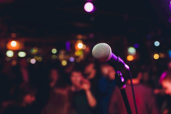 A microphone is in focus on a stand, with a blurred background of colorful lights and people, suggesting a lively event or performance.