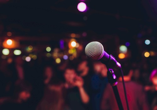 A microphone stands in focus on stage, with a blurred background of colorful lights and a crowd, suggesting a live performance setting.