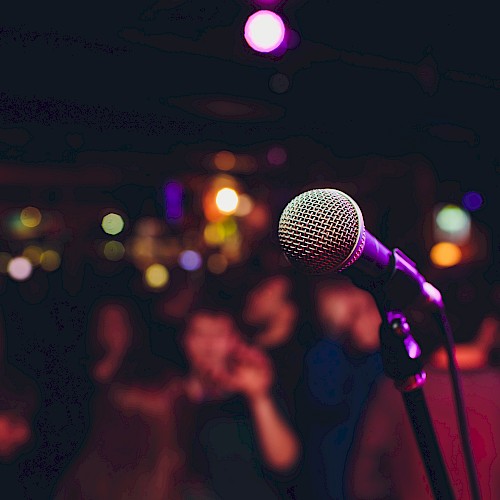 A microphone stands in focus on stage, with a blurred background of colorful lights and a crowd, suggesting a live performance setting.