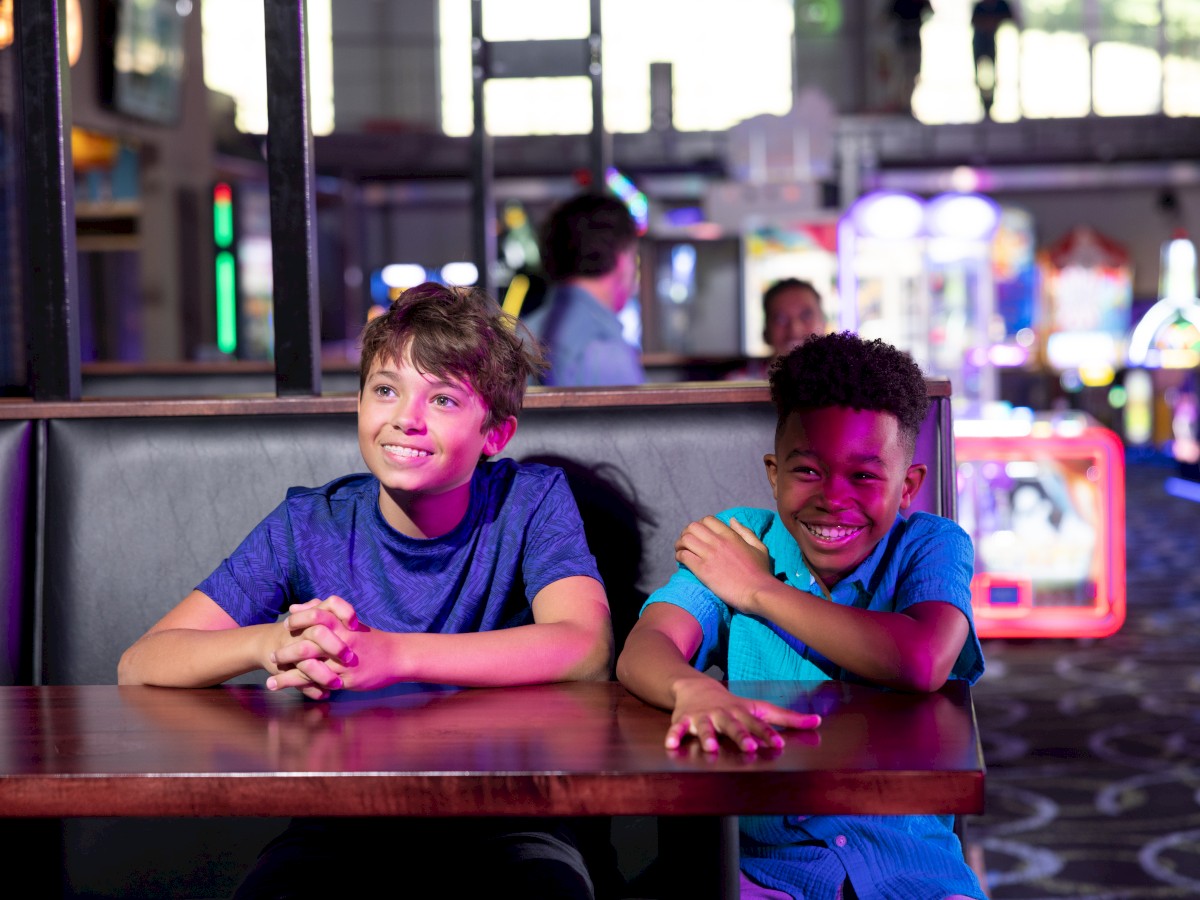 Two kids are sitting at a table in an arcade, smiling and posing for the camera with games in the background.