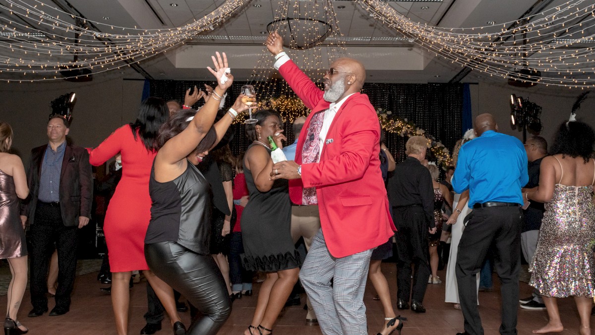 A group of people are dancing energetically at an indoor party under elegant chandeliers. Everyone appears to be enjoying the celebration.