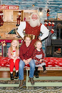 A festive scene with Santa Claus and three children sitting on a bench, surrounded by Christmas decor and gifts in a workshop setting.