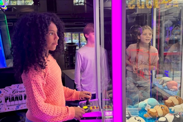 A person is using a claw machine filled with stuffed animals in an arcade, surrounded by colorful lights.