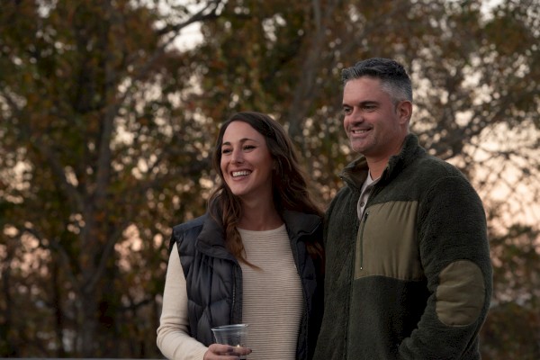 A man and a woman are standing together outdoors, smiling, with trees in the background. She holds a drink in her hand.