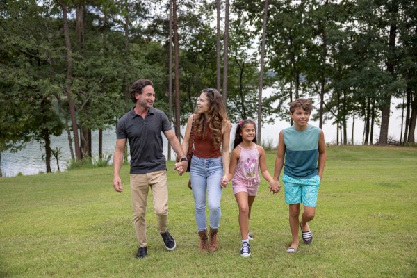 A family of four is walking hand in hand on a grassy area with trees and a lake in the background, enjoying a pleasant day outdoors.