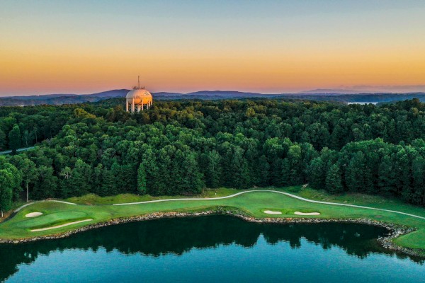 A golf course by a lake at sunset with a domed building and wooded area in the background, under a colorful sky.