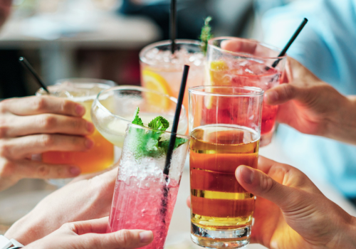 A group of people toasting with assorted colorful drinks, garnished with fruits and herbs, using various glasses and straws.