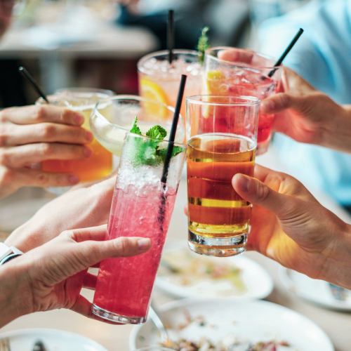 A group of people toasting with assorted colorful drinks, garnished with fruits and herbs, using various glasses and straws.