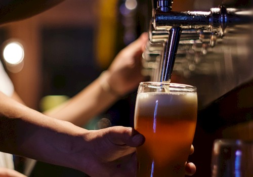 The image shows a person pouring a beer from a tap into a glass at a bar, with a blurred background in warm lighting.