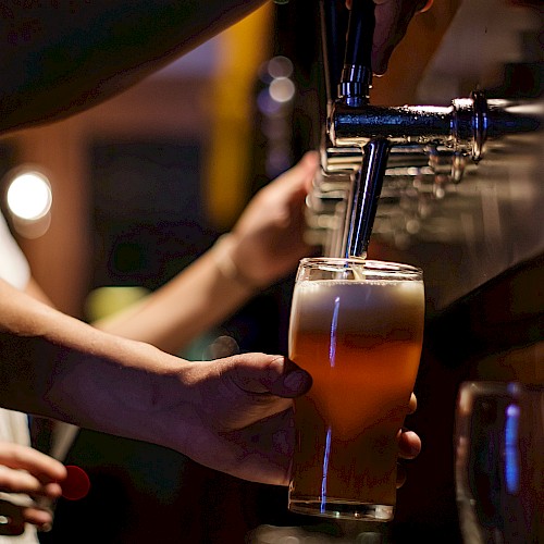 The image shows a person pouring a beer from a tap into a glass at a bar, with a blurred background in warm lighting.