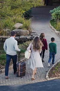 A group of four people walking on a path with luggage, surrounded by landscaped greenery and leading to a building.