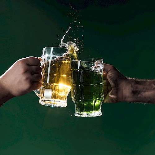 Two hands clinking beer mugs with beer splashing, set against a dark green background, creating a dynamic celebratory moment.