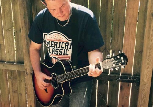 A person is playing an acoustic guitar while standing in front of a wooden fence, wearing a t-shirt and jeans.